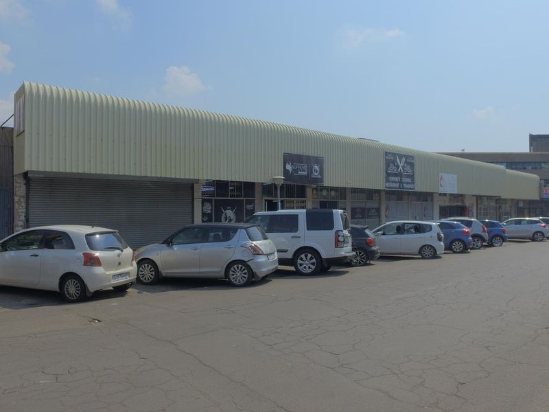 Front exterior view of a commercial retail strip center featuring a corrugated metal roof, roller shutter doors, and an adjacent paved parking area.