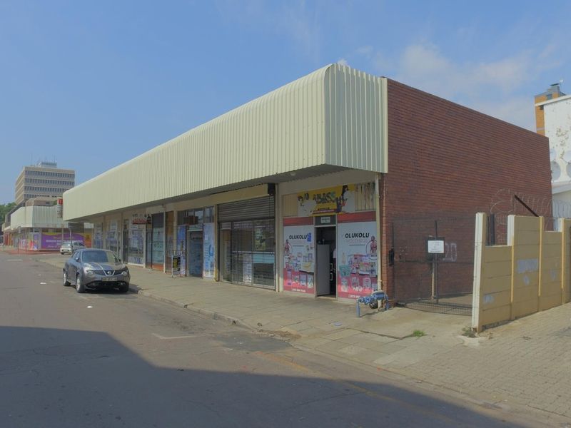 Street-level view of a retail property featuring a brick facade, metal awning, and security roller shutters along a paved sidewalk.
