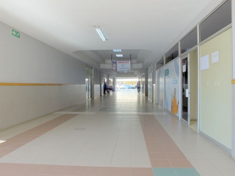 A bright, tiled commercial hallway featuring glass storefront partitions and overhead fluorescent lighting, providing access to various retail or office units.