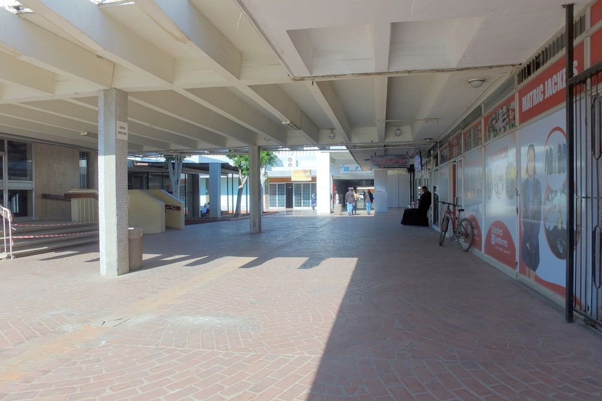 A covered outdoor walkway area in a retail complex featuring brick paving, concrete structural pillars, and storefront access points.