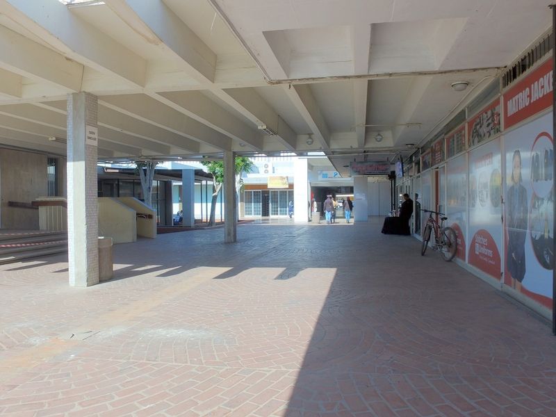 A covered outdoor walkway area in a retail complex featuring brick paving, concrete structural pillars, and storefront access points.