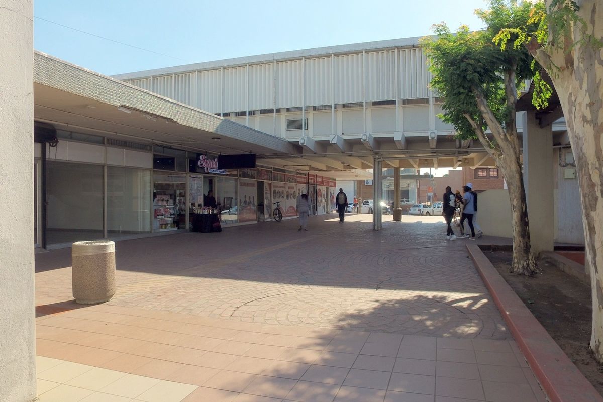 A view of a retail center walkway featuring paved surfaces, concrete structural pillars, and multiple storefront glass entrances under a covered canopy.