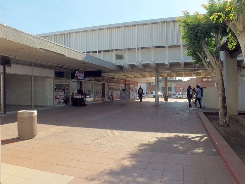 A view of a retail center walkway featuring paved surfaces, concrete structural pillars, and multiple storefront glass entrances under a covered canopy.