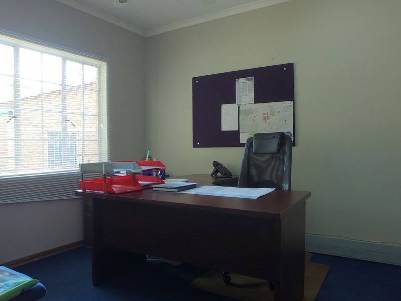 A functional office space featuring carpeted flooring, a large window with fitted blinds, and neutral wall finishes.