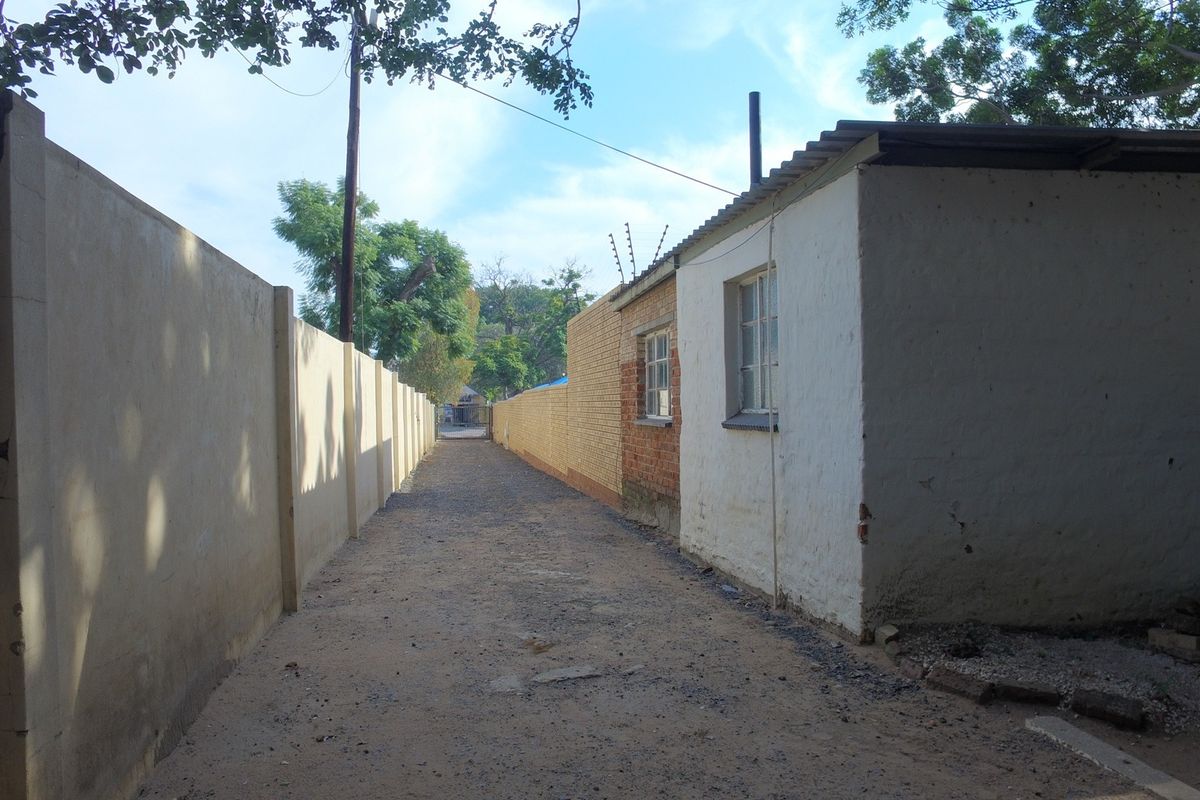 A narrow exterior rear alleyway featuring a perimeter boundary wall with electric fencing and a basic structure with corrugated roofing.