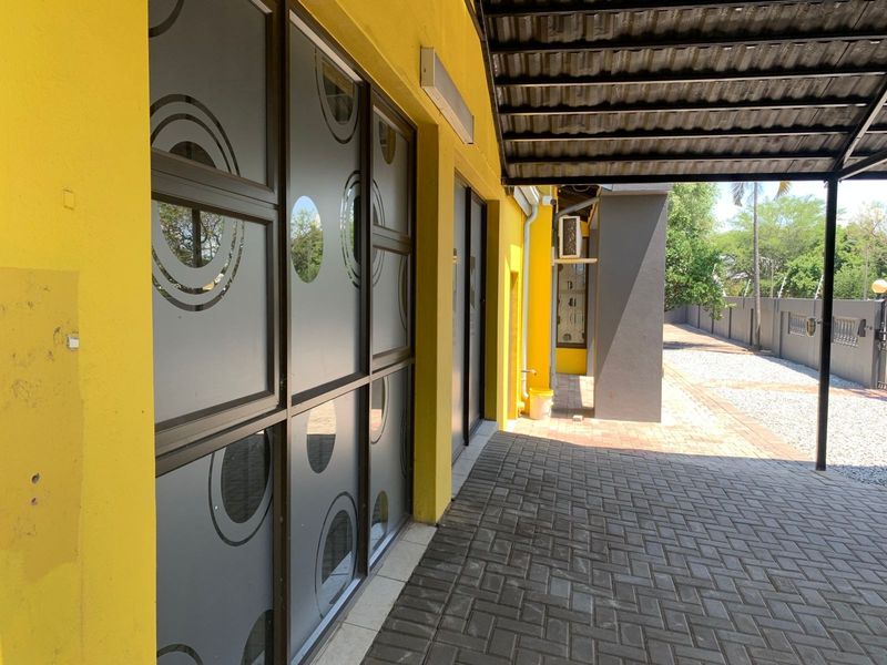 A covered paved walkway along the side of a yellow office building featuring frosted glass windows and metal roofing.