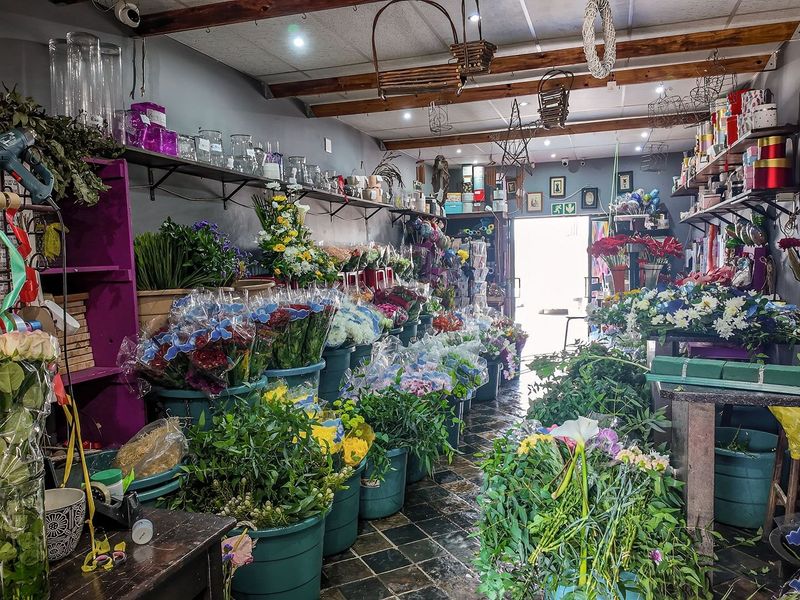 A commercial workspace currently utilized as a floral shop, featuring exposed timber ceiling beams, wall-mounted shelving, and durable tiled flooring.