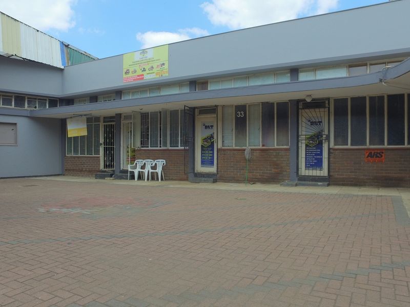 Rear exterior view of a commercial retail property featuring brick paving, multiple entrance points with security gates, and large windows.