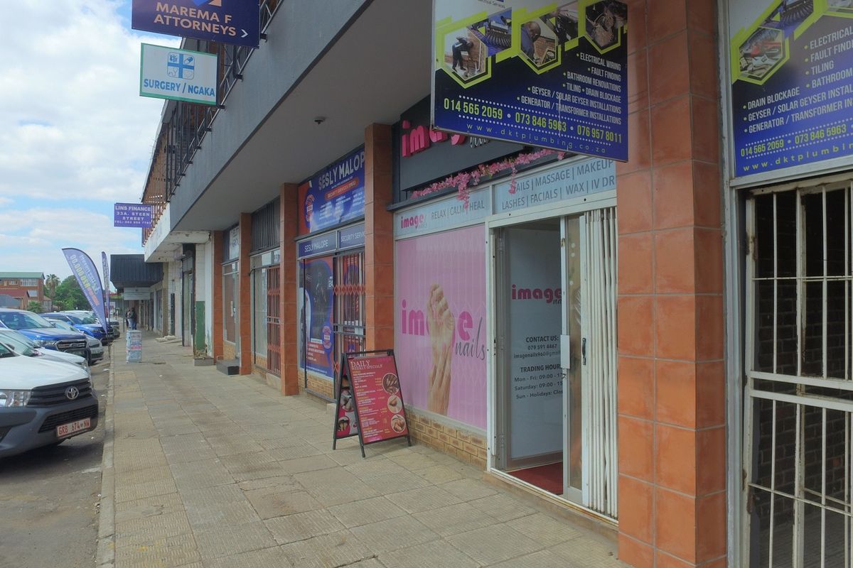 Street-level view of a retail commercial building featuring multiple storefronts with signage, security gates, and a paved pedestrian walkway.