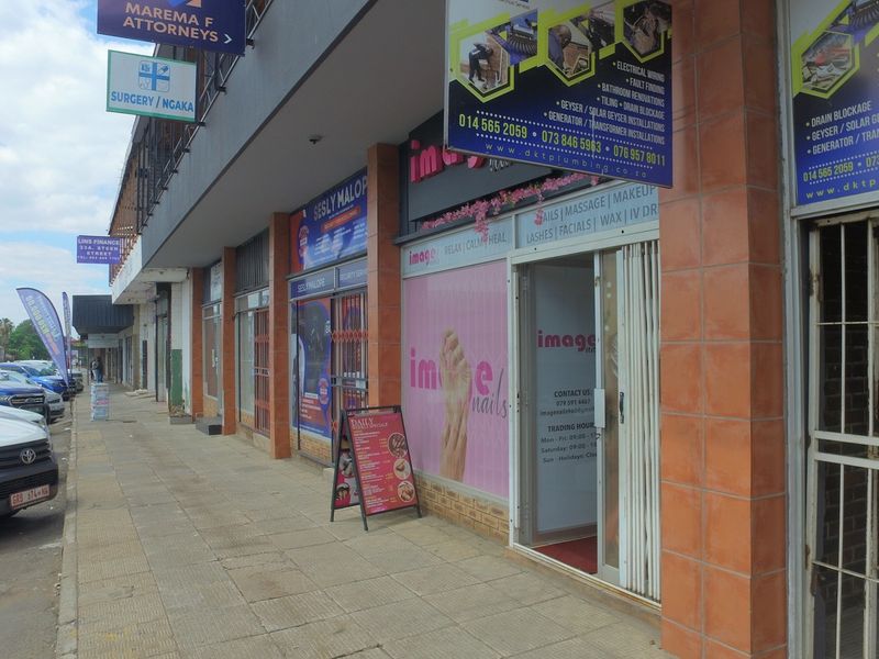 Street-level view of a retail commercial building featuring multiple storefronts with signage, security gates, and a paved pedestrian walkway.