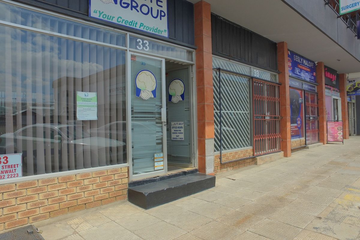 Street-level view of a retail storefront featuring a glass entrance, brick facade, and security gate, located along a paved sidewalk.