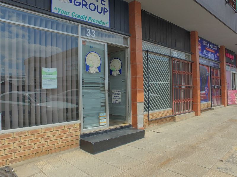 Street-level view of a retail storefront featuring a glass entrance, brick facade, and security gate, located along a paved sidewalk.