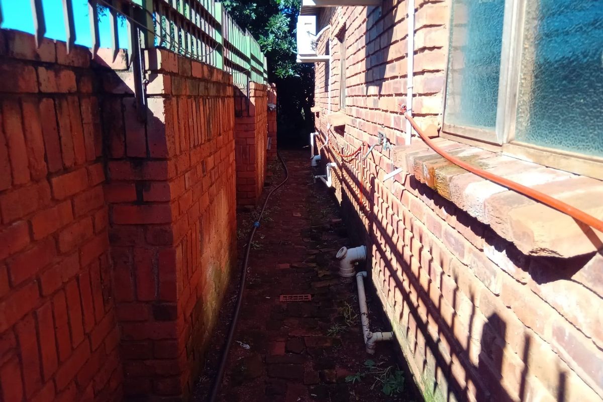 A narrow side passage featuring brick boundary walls, paved flooring, and exposed external utility infrastructure.