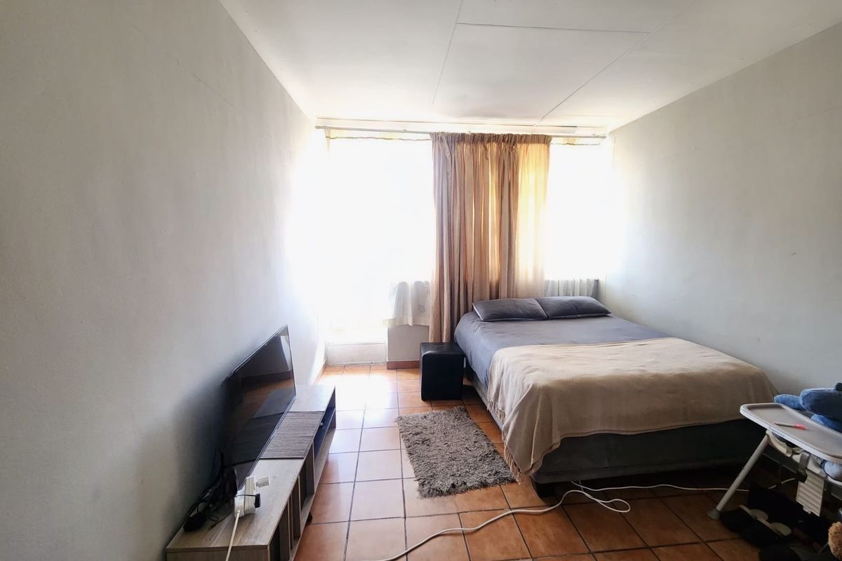 A compact bedroom featuring tiled flooring and a large window, providing natural light. The space requires cosmetic updates to improve its overall condition.
