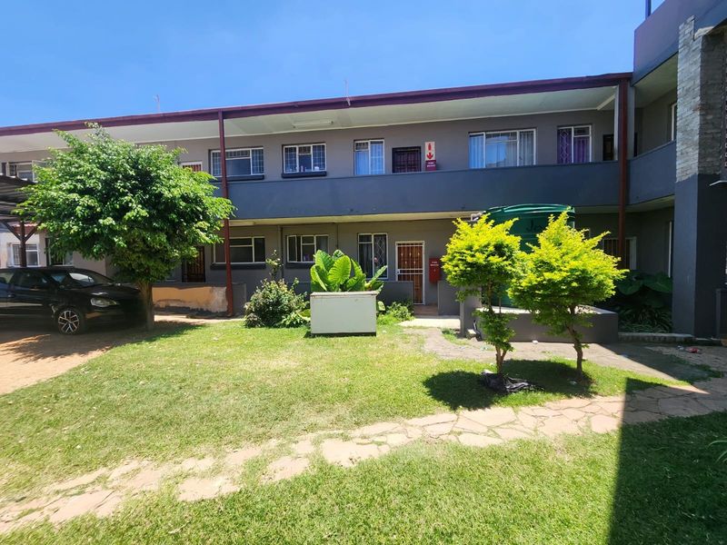 Exterior view of an apartment complex featuring a shared garden area, paved walkways, and visible security features including security gates and fire safety equipment.