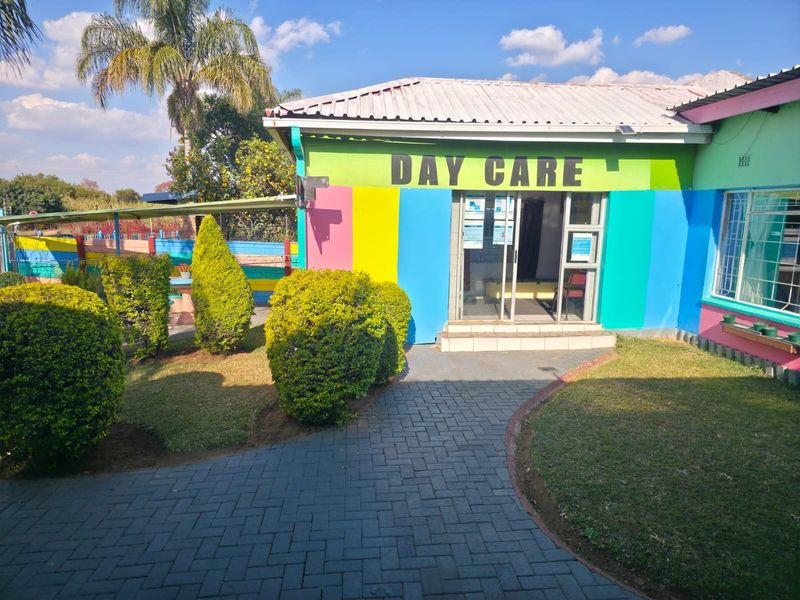 Front entrance of a day care facility featuring a paved walkway, landscaped garden, and colorful exterior facade with sliding glass door access.
