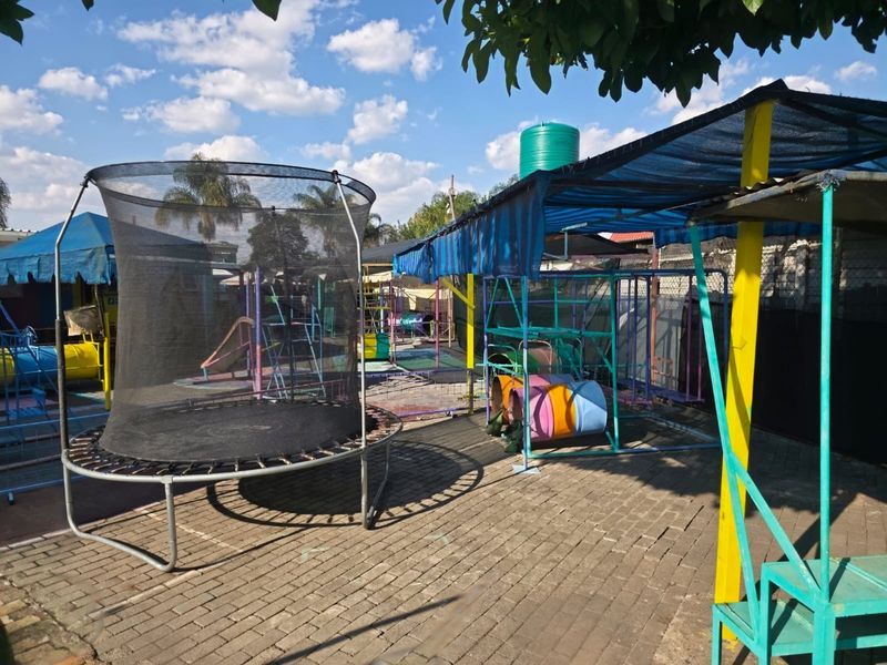 Outdoor recreational area featuring a paved courtyard, shade cloth structures, and various play equipment, suitable for a childcare or sports facility.