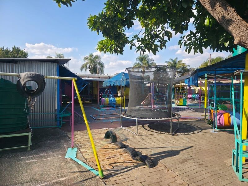 Outdoor play area featuring a paved courtyard, corrugated metal structures, and various fixed shade installations within a commercial property setting.