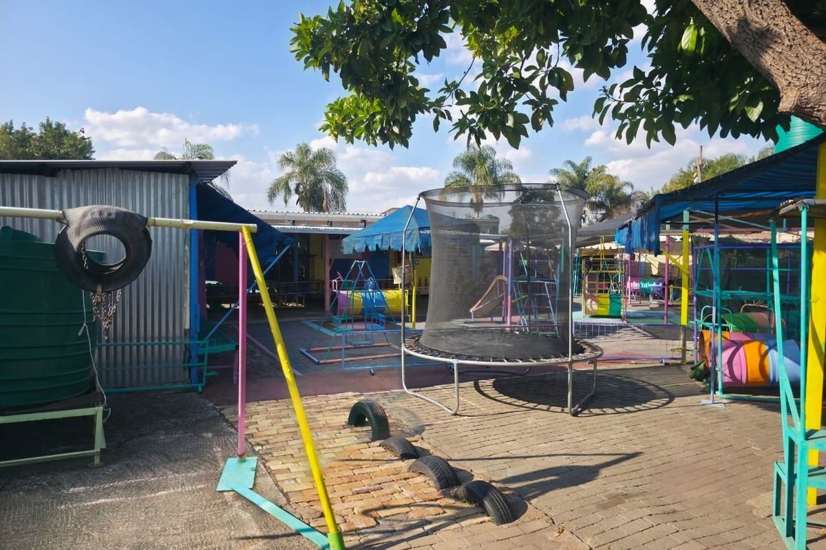Outdoor play area featuring a paved courtyard, corrugated metal structures, and various fixed shade installations within a commercial property setting.