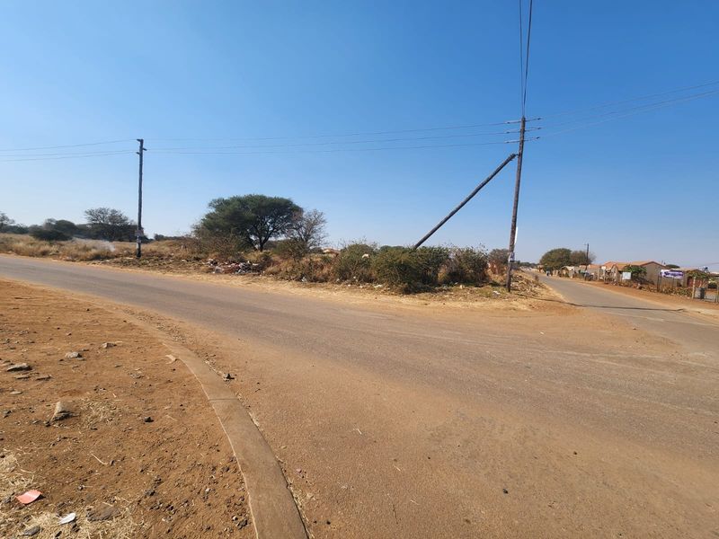 A street-level view of a vacant commercial land plot situated at a road intersection, featuring existing utility infrastructure and surrounding natural landscape.