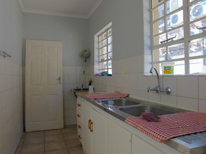 A functional laundry room featuring a double stainless steel sink, tiled splashbacks, built-in cabinetry, and tiled flooring with natural light from windows.