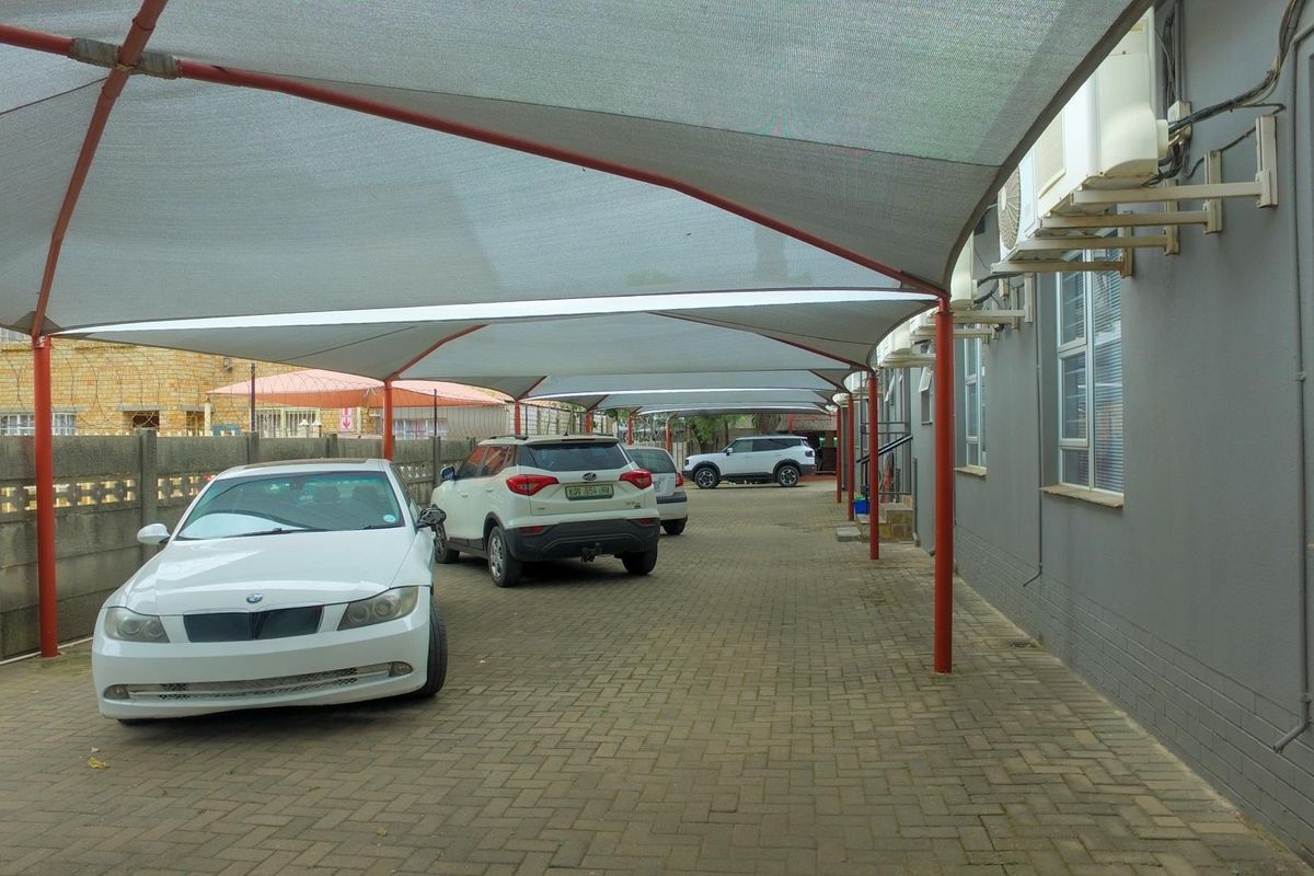 A paved parking area featuring a large shade cloth carport structure, external air conditioning units, and secure perimeter walling.
