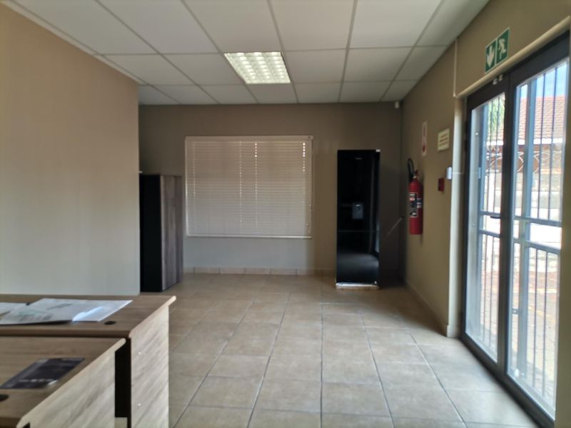 A functional office space featuring tiled flooring, a suspended ceiling with recessed lighting, and direct access to the exterior via a secure glass door.