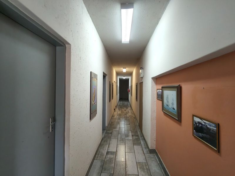 A long, functional hallway featuring tiled flooring, multiple access doors, and overhead fluorescent lighting, providing access to various rooms within the guest house.