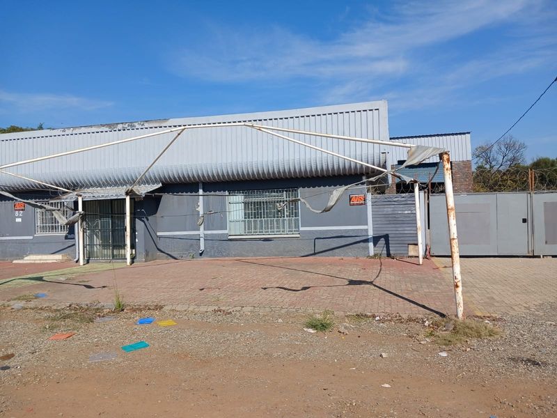 Industrial property exterior featuring a paved frontage, security bars on windows, and a damaged metal frame structure with perimeter fencing.
