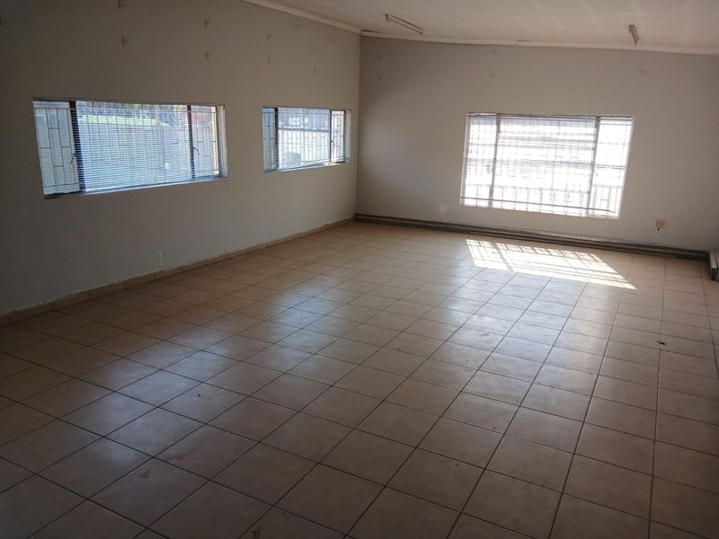 An empty, open-plan office space featuring tiled flooring, multiple windows with security bars, and basic fluorescent lighting.