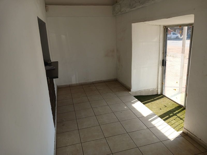 A small, empty industrial entry space featuring tiled flooring, a glass entrance door, and a built-in service counter, requiring minor cosmetic wall repairs.
