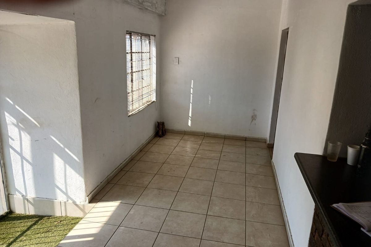 An empty, sunlit interior space featuring tiled flooring, security bars on the window, and a built-in counter, suitable for a living or utility area.