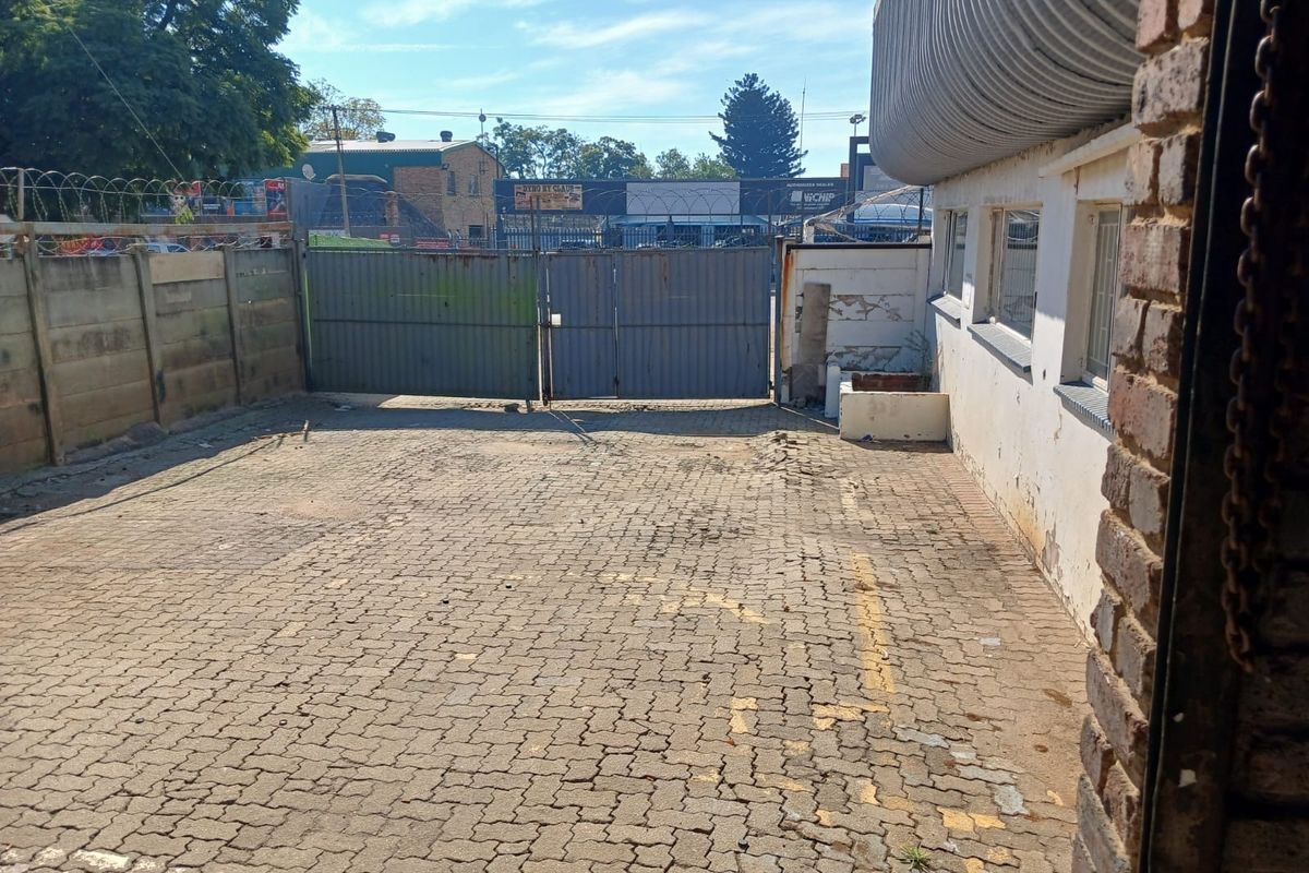 A paved industrial yard area featuring a metal security gate, perimeter wall with razor wire, and brick building facade with corrugated roof detailing.