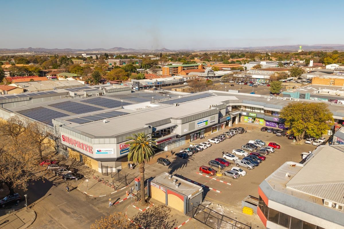 Aerial view of a commercial shopping center featuring a large roof with solar panels, an adjacent parking lot, and surrounding urban infrastructure.