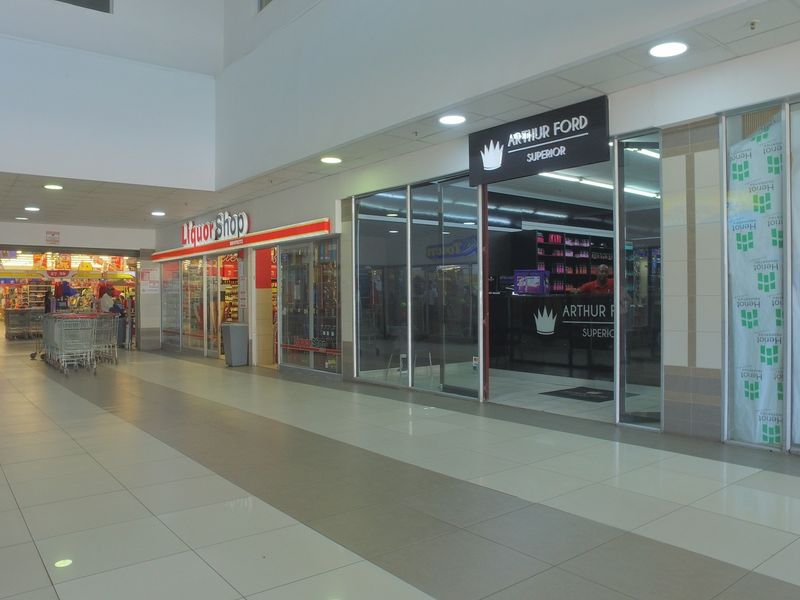 A well-maintained commercial shopping center hallway featuring tiled floors, recessed lighting, and multiple retail storefronts with glass facades.