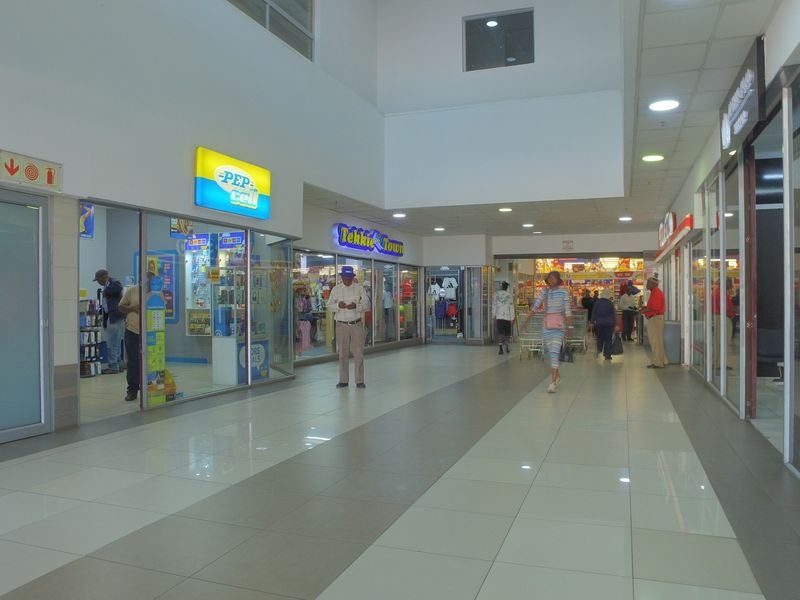 A view of a commercial shopping center corridor featuring tiled flooring, recessed lighting, and multiple glass-fronted retail storefronts.