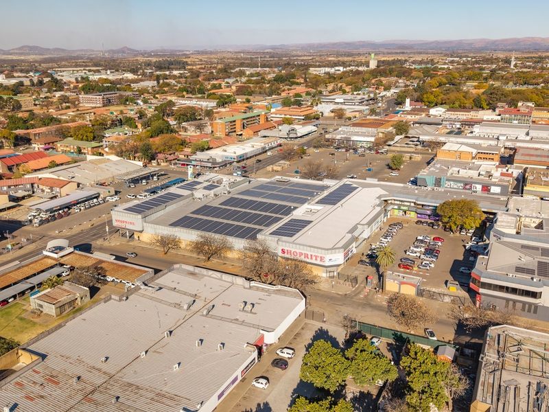 Aerial view of a commercial shopping center featuring a large roof-mounted solar panel array and adjacent paved parking facilities within an urban setting.