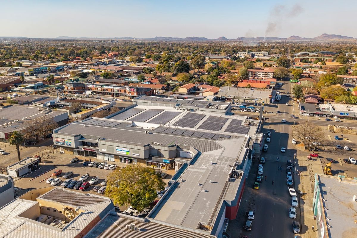 Aerial view of a commercial shopping center featuring extensive rooftop solar panel installations and an adjacent parking area within a busy urban environment.