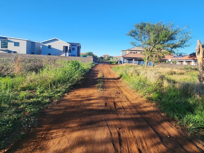 A view of an unpaved dirt road leading through a developing residential area with surrounding houses.