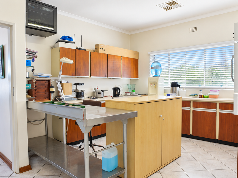A functional office space featuring built-in wooden cabinetry, a stainless steel workstation, tiled flooring, and large windows with venetian blinds.
