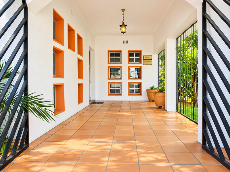 A well-maintained covered entrance area featuring terracotta tiled flooring, decorative orange-framed wall niches, and secure metal gate access.