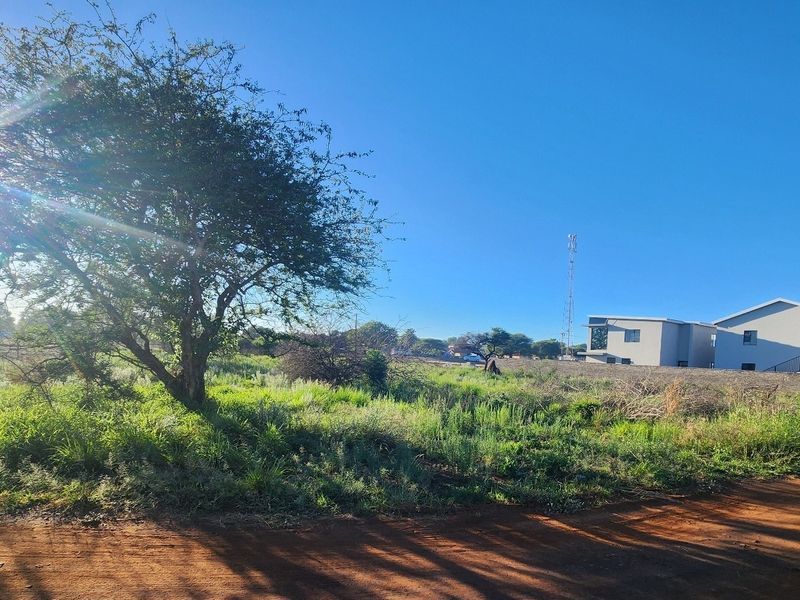 A view of the vacant commercial land featuring natural vegetation, dirt road access, and perimeter fencing, with neighboring structures visible in the background.
