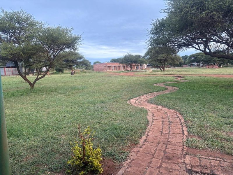 A spacious garden area featuring a winding brick pathway leading across a large lawn with mature trees and distant building structures.