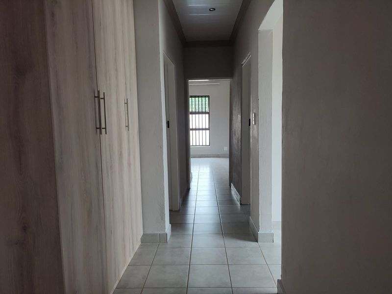 A clean, well-lit hallway featuring neutral tiled flooring and extensive built-in wooden cabinetry for storage, leading towards a bright room with security bars.