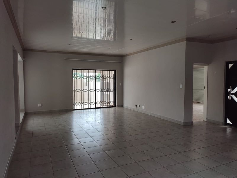 A spacious, empty living area featuring neutral tiled flooring, recessed downlighting, and a sliding glass door with a security gate leading outdoors.