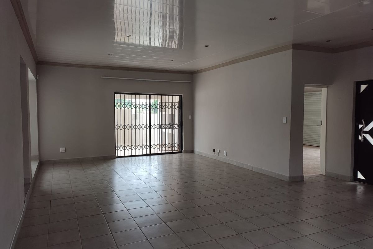 A spacious, empty living area featuring neutral tiled flooring, recessed downlighting, and a sliding glass door with a security gate leading outdoors.