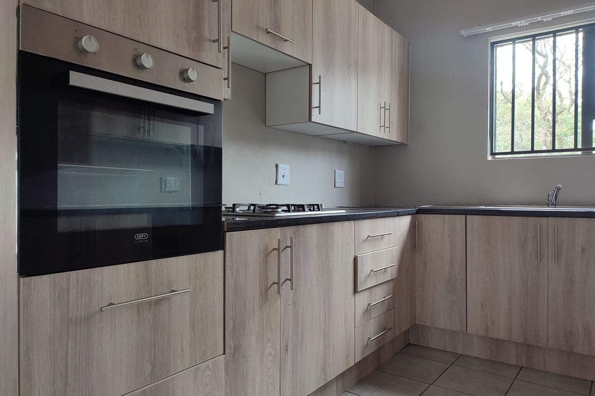 A functional kitchen featuring light wood-look cabinetry, built-in oven, gas hob, dark countertops, and tiled flooring with a window fitted with security bars.