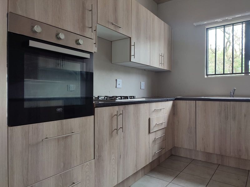 A functional kitchen featuring light wood-look cabinetry, built-in oven, gas hob, dark countertops, and tiled flooring with a window fitted with security bars.