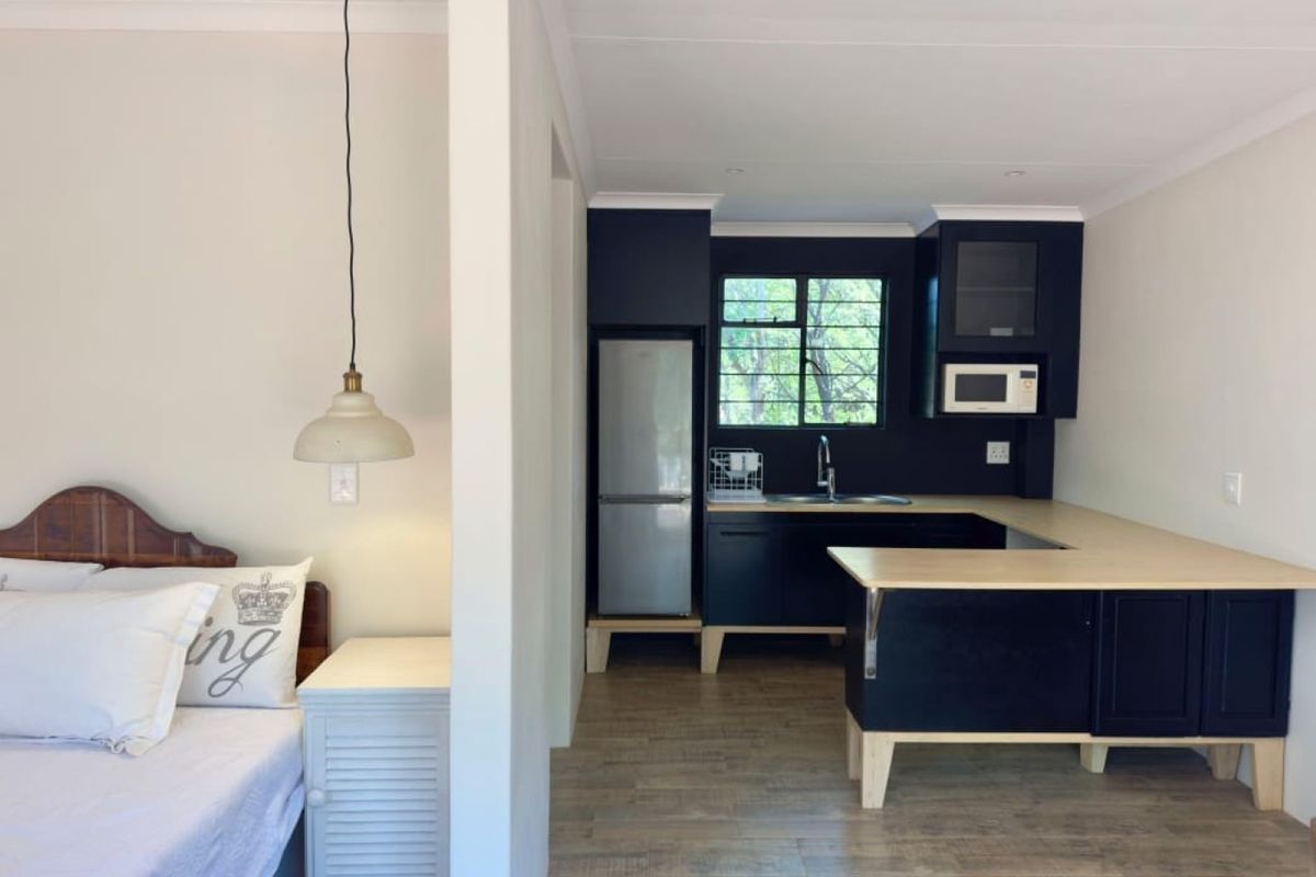 Open-plan studio layout featuring a modern kitchen with dark cabinetry, light wood-look countertops, and an adjacent bedroom area with a pendant light.