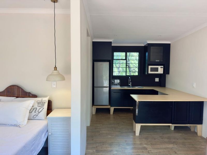 Open-plan studio layout featuring a modern kitchen with dark cabinetry, light wood-look countertops, and an adjacent bedroom area with a pendant light.